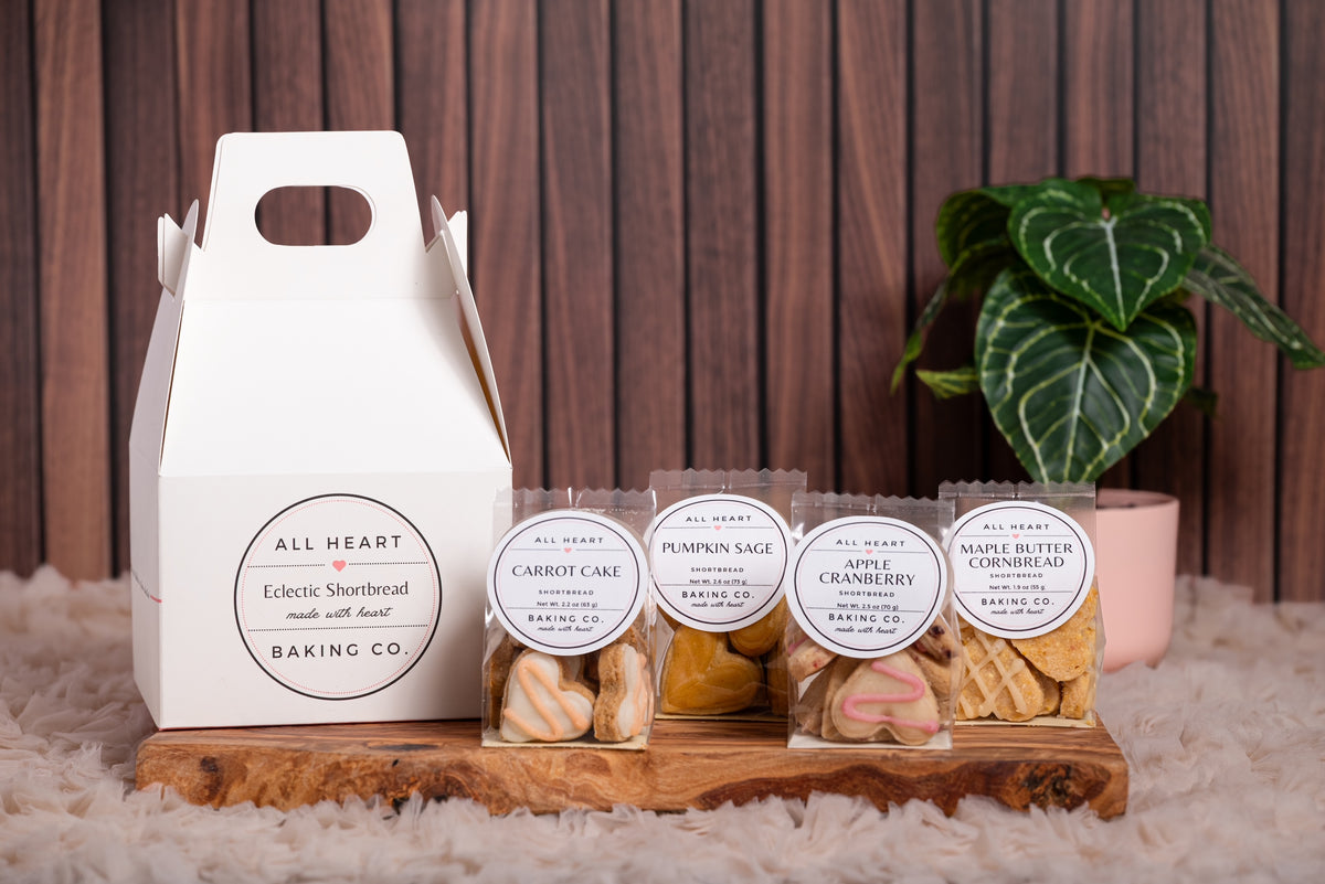 A white paper bakery gift box labeled 'All Heart Baking Co' stands on a wooden tray beside four individually wrapped bags of miniature heart-shaped shortbread cookies with matching round labels identifying the flavors as carrot cake, pumpkin sage, apple cranberry, maple butter cornbread. The scene is styled on a white faux-fur surface against a dark wood plank background, with a green potted plant behind.