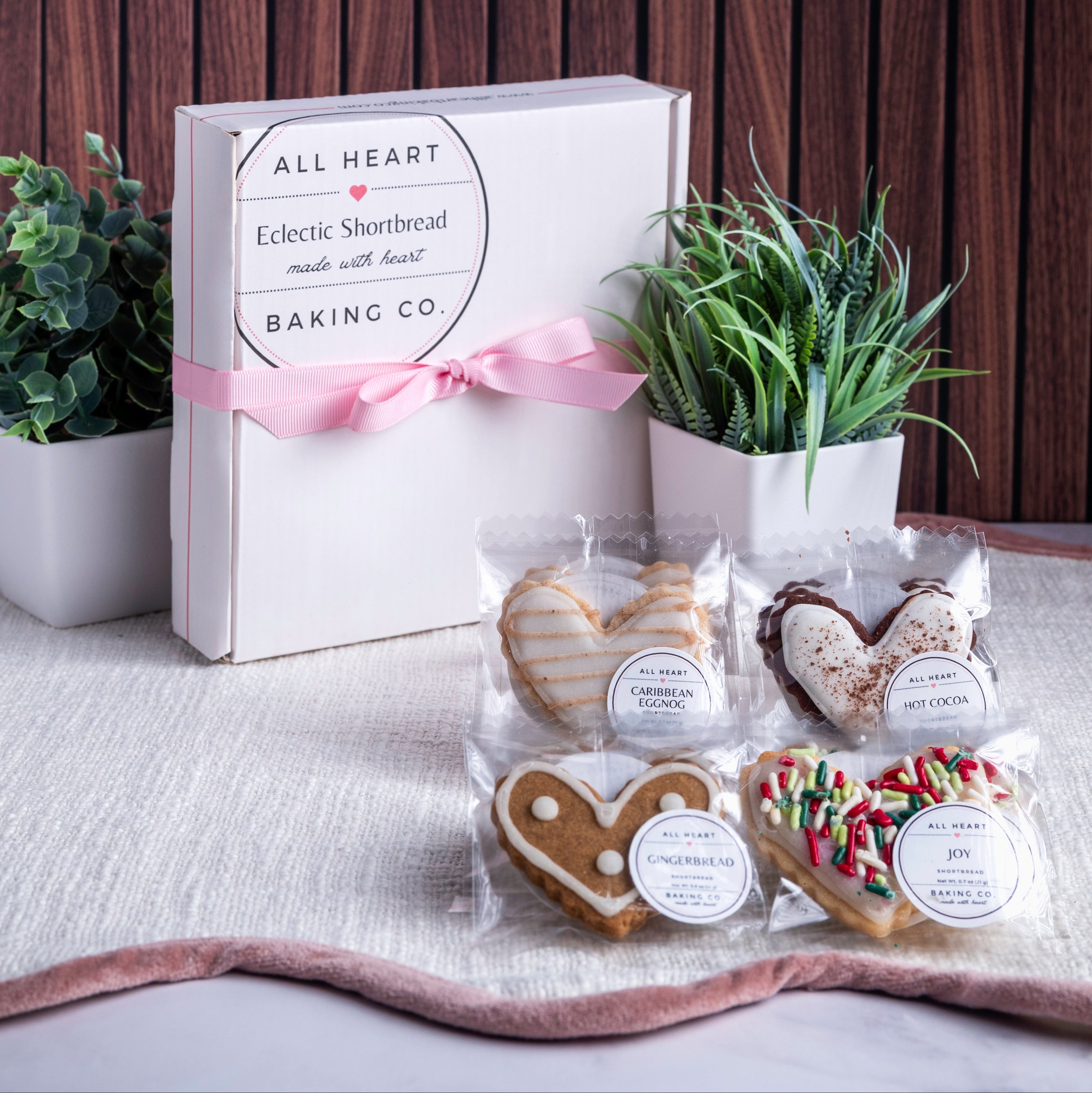 Heart-shaped cookies in clear packaging with a 'All Heart Baking Co.' box and plants in the background.